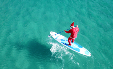 Père noël au milieu de la mer sur une planche de surf.