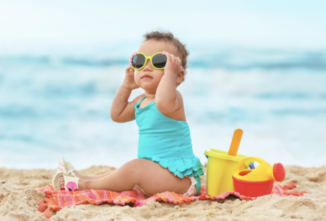 Bébé à la plage avec des lunettes de soleil et des jouets de plage à côté.