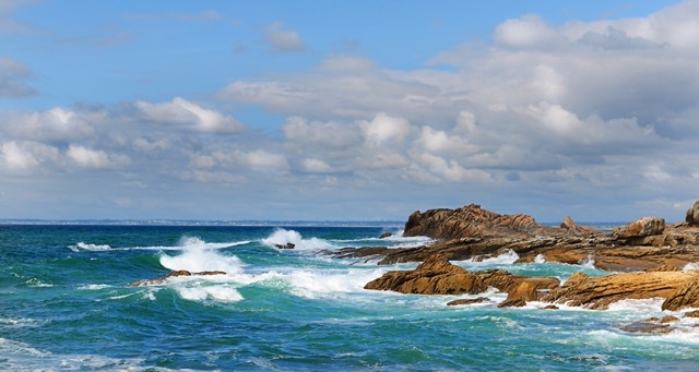 Une côte rocheuse avec un ciel en fond plein de nuages. Mais il fait beau. La mer, d'un joli bleu est vive et des vagues viennent rompre contre les rochers. 